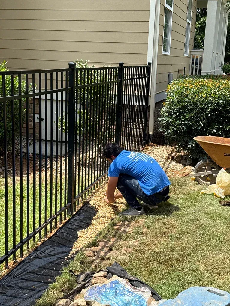 Person tending to gravel border next to black metal fence and house.