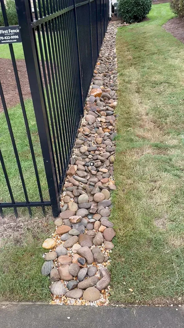 Stone-lined drainage ditch next to a black metal fence and lawn.