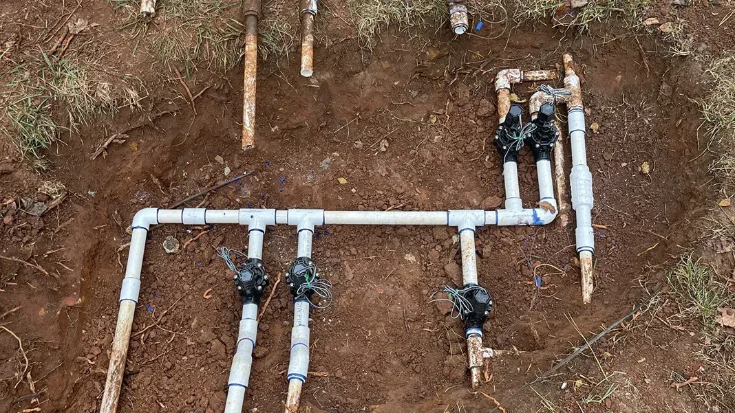 Pipes in a small excavated hole in grassy ground, likely plumbing repair. Brown shoes on the bottom left.