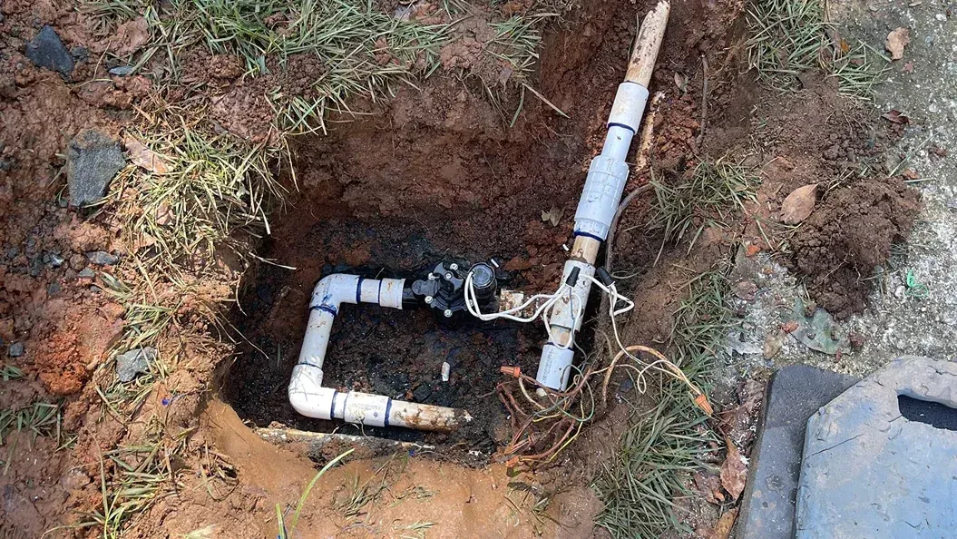 Irrigation system valve in a dirt hole with white PVC pipes and electrical wiring.
