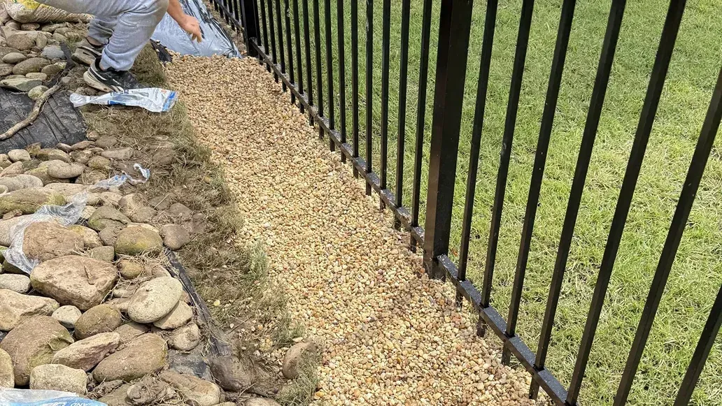 Person spreading wood pellets along a black fence, beside a rock border and lawn.
