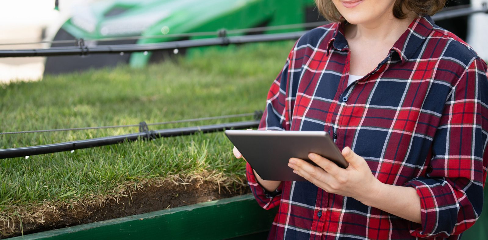 Woman in plaid shirt using a tablet in a field of grass next to farming equipment.
