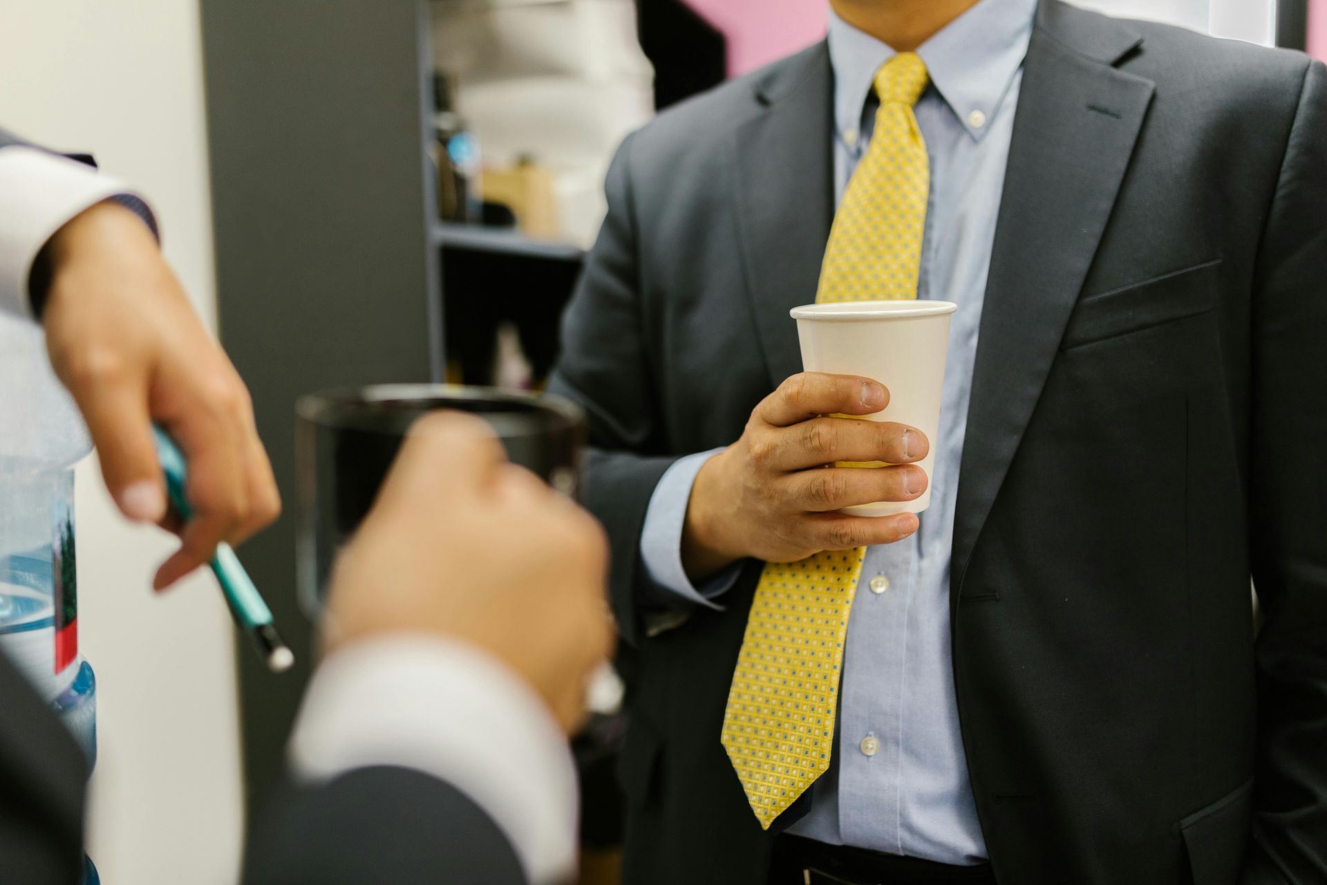 Man in suit holds coffee cup, talking to another man holding a pen.