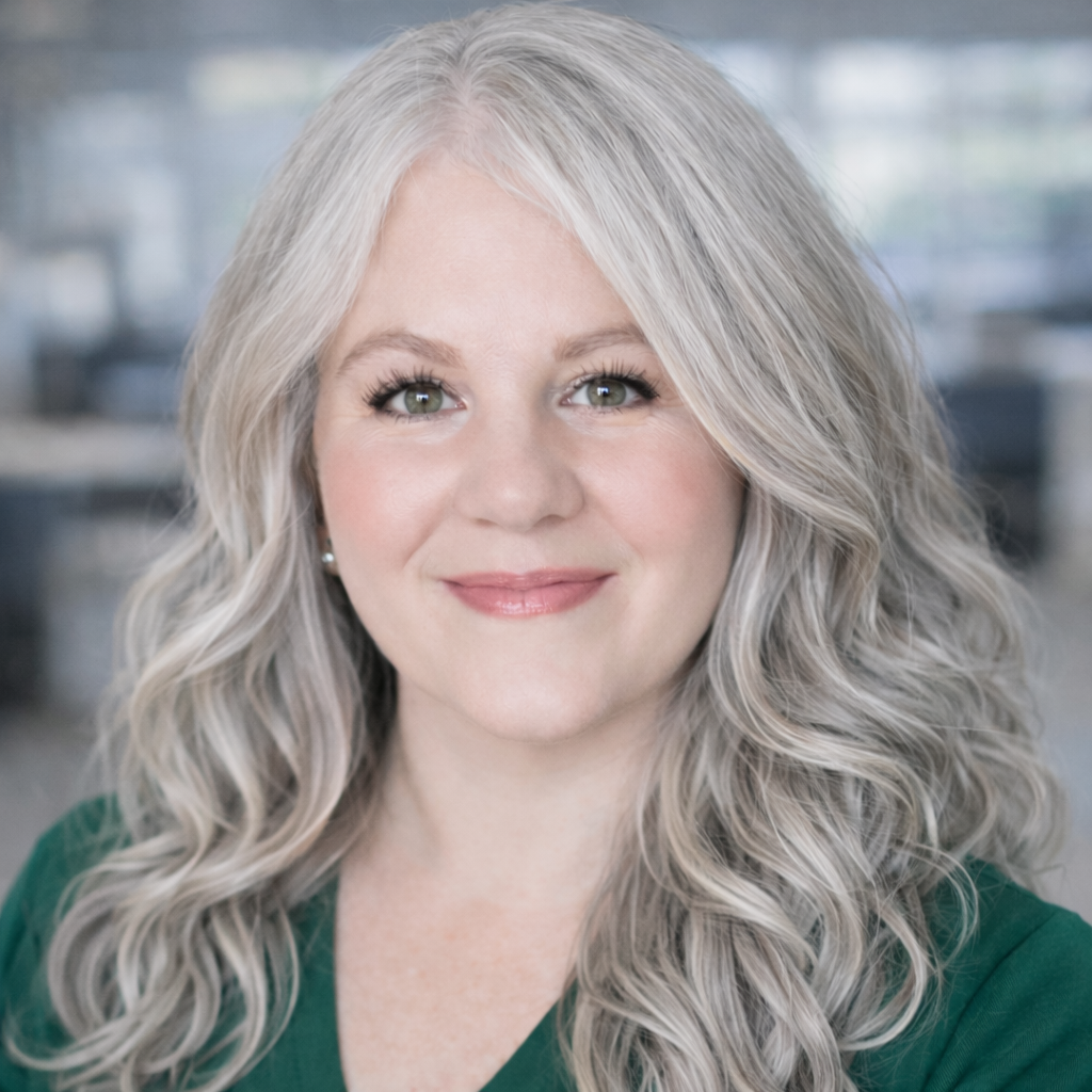 Woman with long, wavy gray hair smiles, wearing a green shirt, soft makeup in an office setting.
