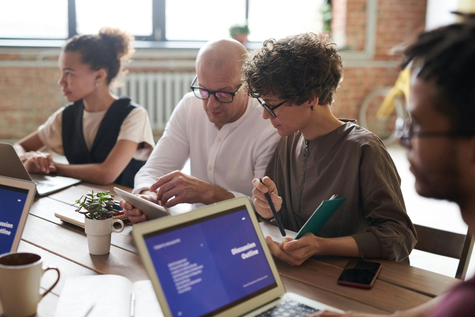 People in an office at a wooden table, working on laptops and tablets, discussing documents.