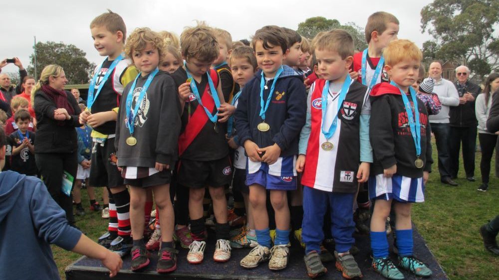 Children in sports uniforms standing on a podium wearing participation medals during an outdoor event.