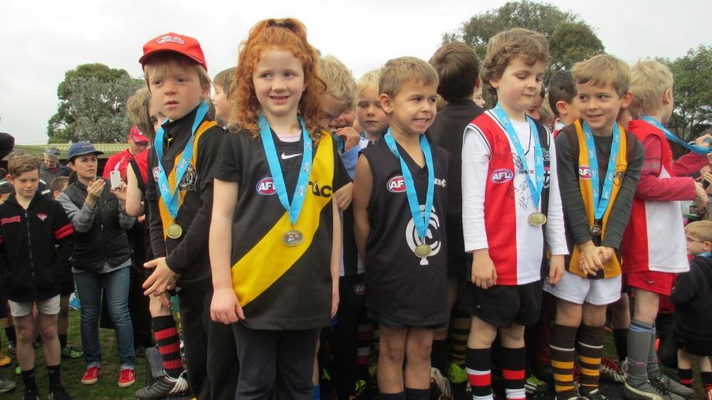 Children in various team uniforms stand in a row wearing medals at an outdoor sports event.