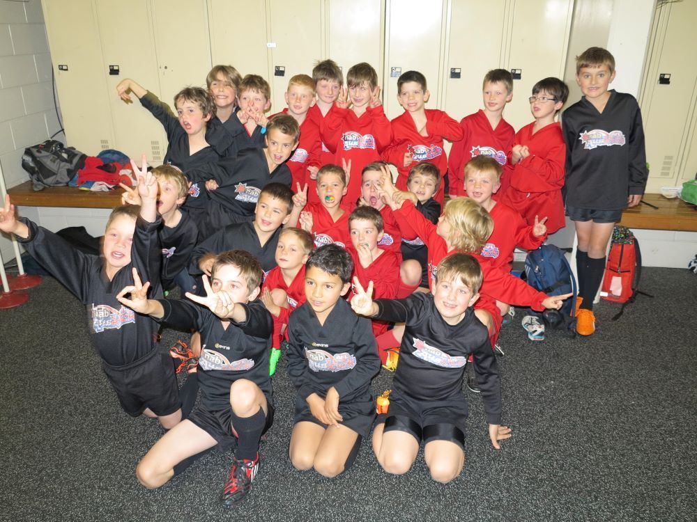 A group of young soccer players in black and red team uniforms pose together in a locker room with lockers in the back.