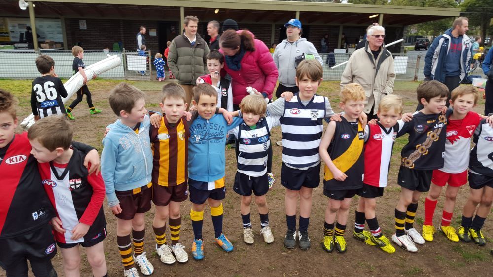 A row of children in various Australian rules football uniforms standing on a field with adults in the background.