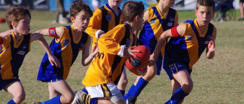 Children in yellow and blue uniforms playing Australian rules football on a grass field, one holding the ball.