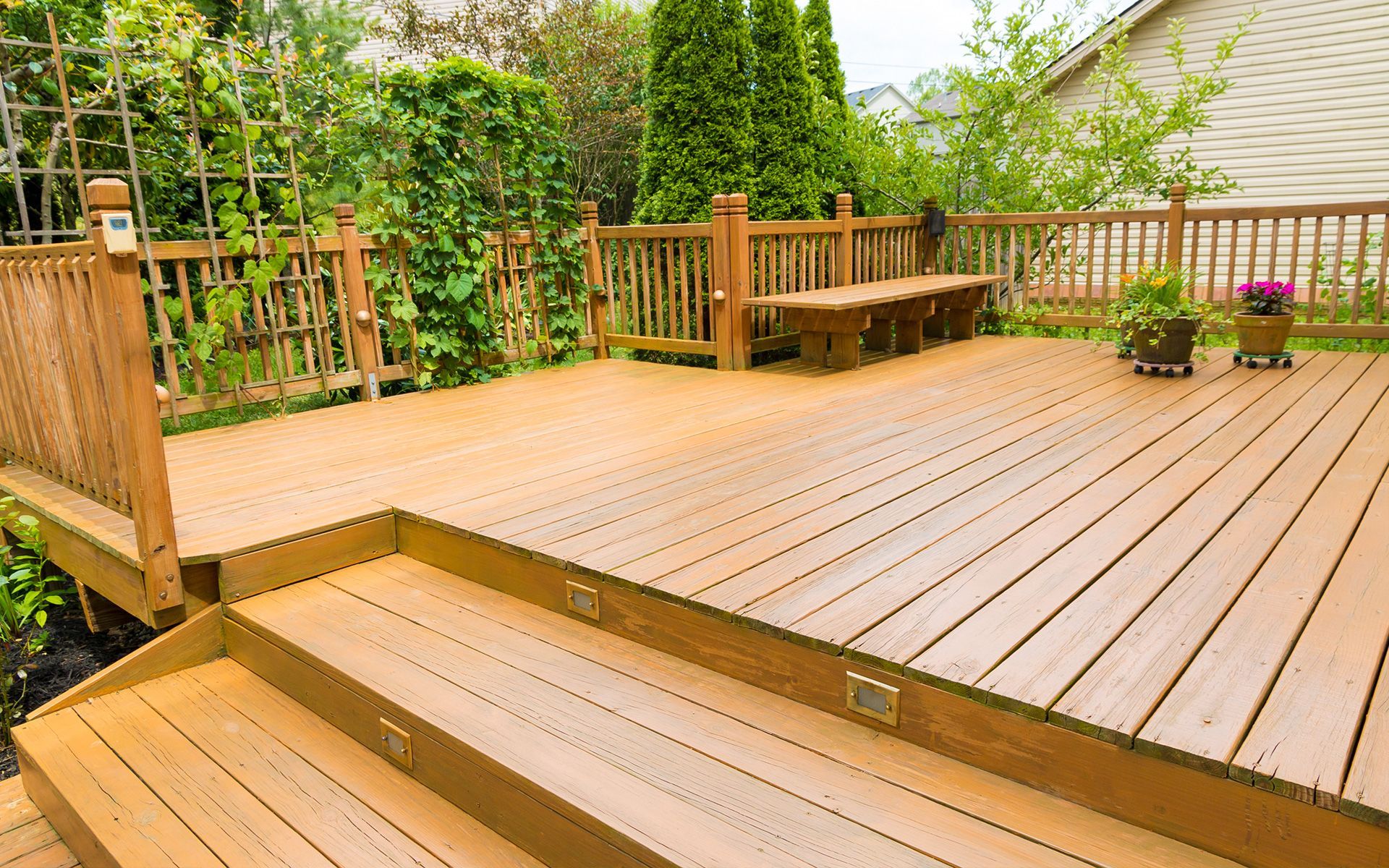 Wooden deck with built-in bench, steps, and railing; surrounded by greenery and a house.