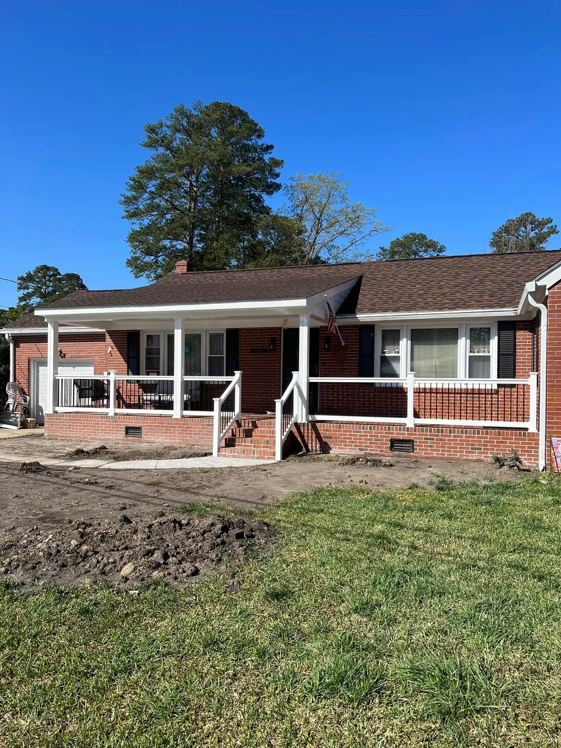 Brick house with white porch and railing, brown roof, and small yard on a sunny day.