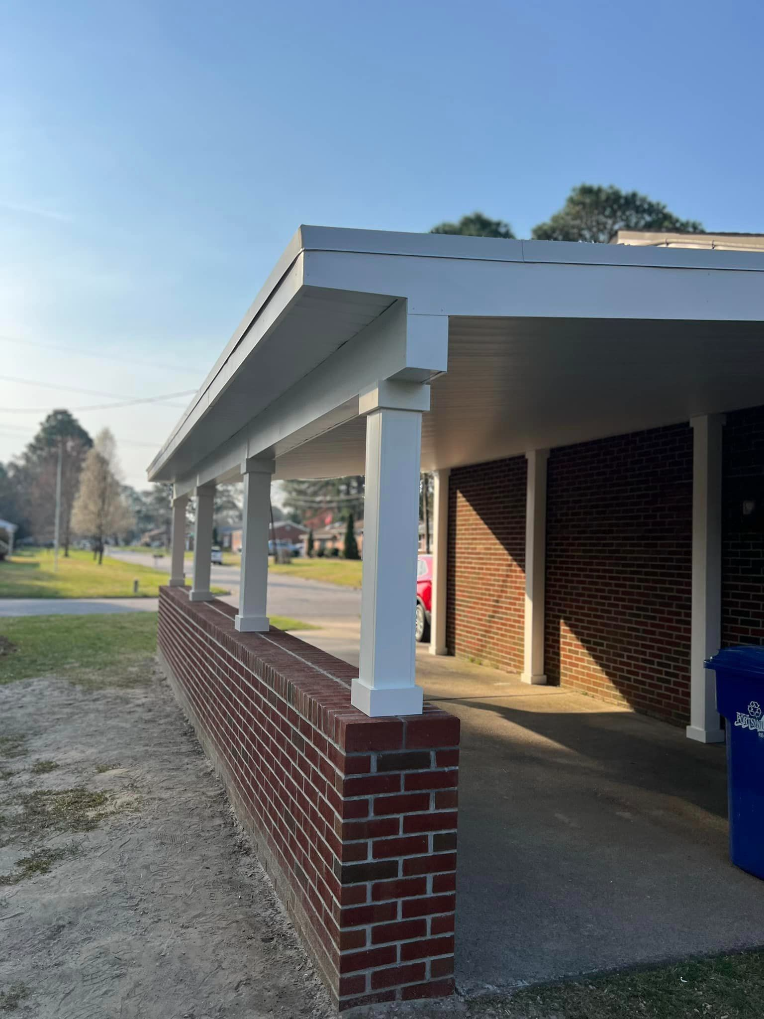 Brick carport with white columns and roof, fronted by a brick wall, overlooking a street on a sunny day.