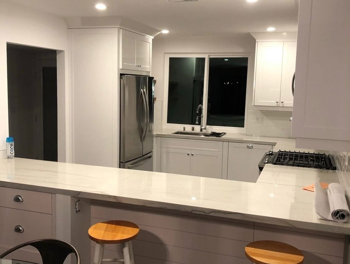 Bright modern kitchen with white cabinets, stainless fridge, island, and two stools at night