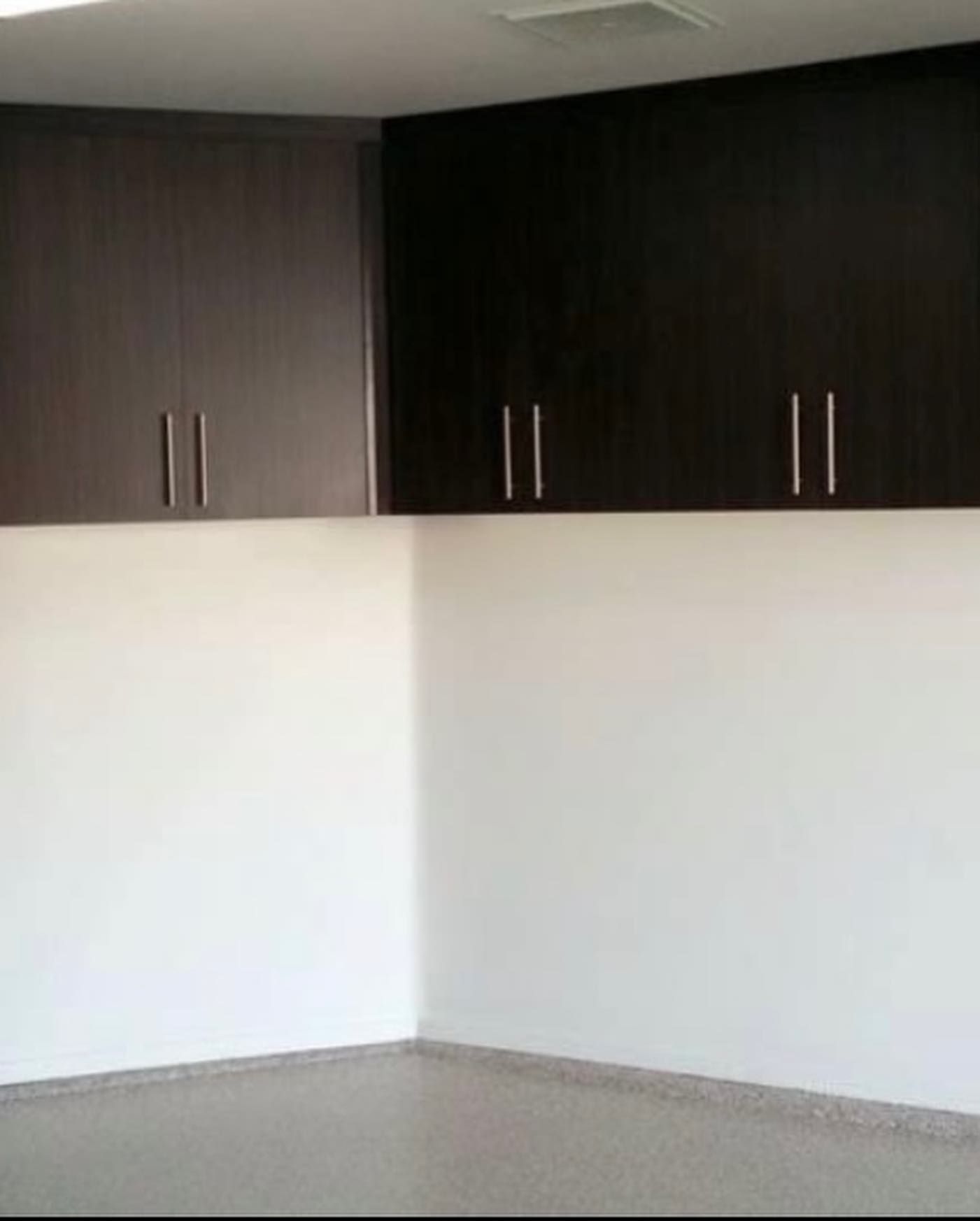 Empty room corner with white walls, gray floor, and dark wall cabinets above.