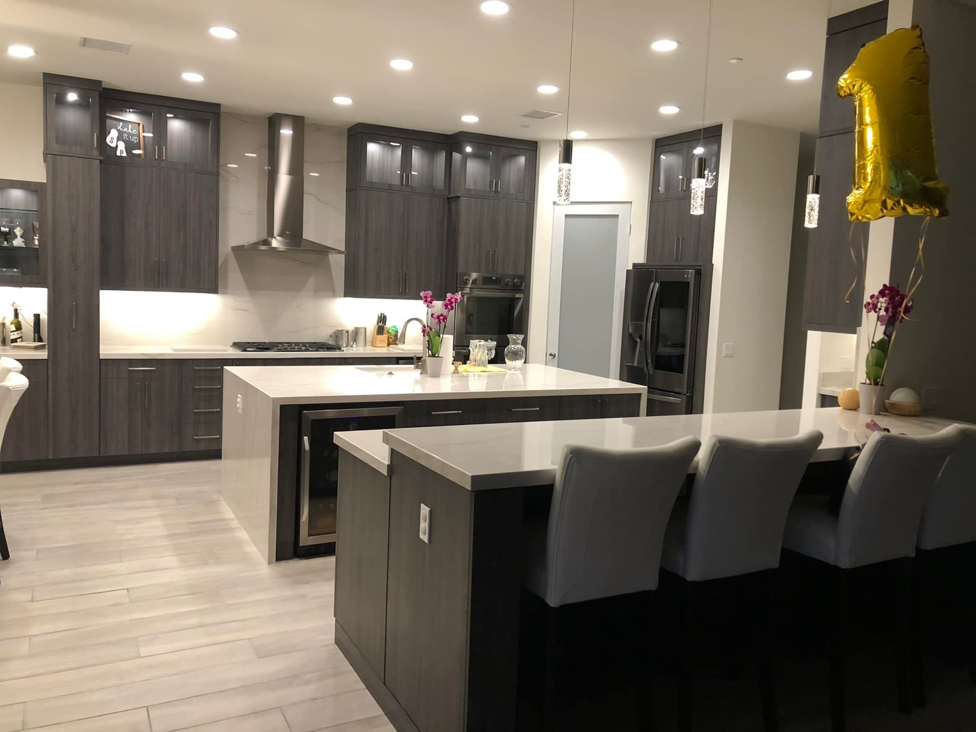 Modern kitchen and dining area with dark cabinets, white countertops, and bar stools under recessed lights