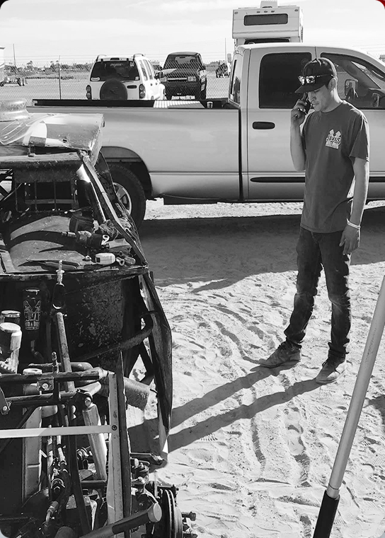 Man on phone by a modified vehicle; truck, RV, and other vehicles in the background. Sandy ground, sunny day.