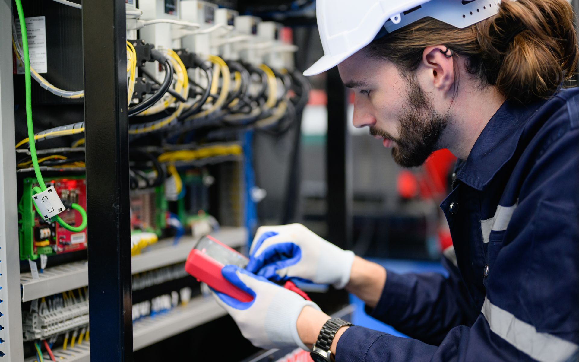 Electrician in white hard hat and gloves tests electrical panel with multimeter.