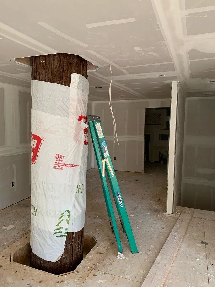 A large tree trunk extends from the floor through a ceiling. A green ladder leans against it inside a room under construction.