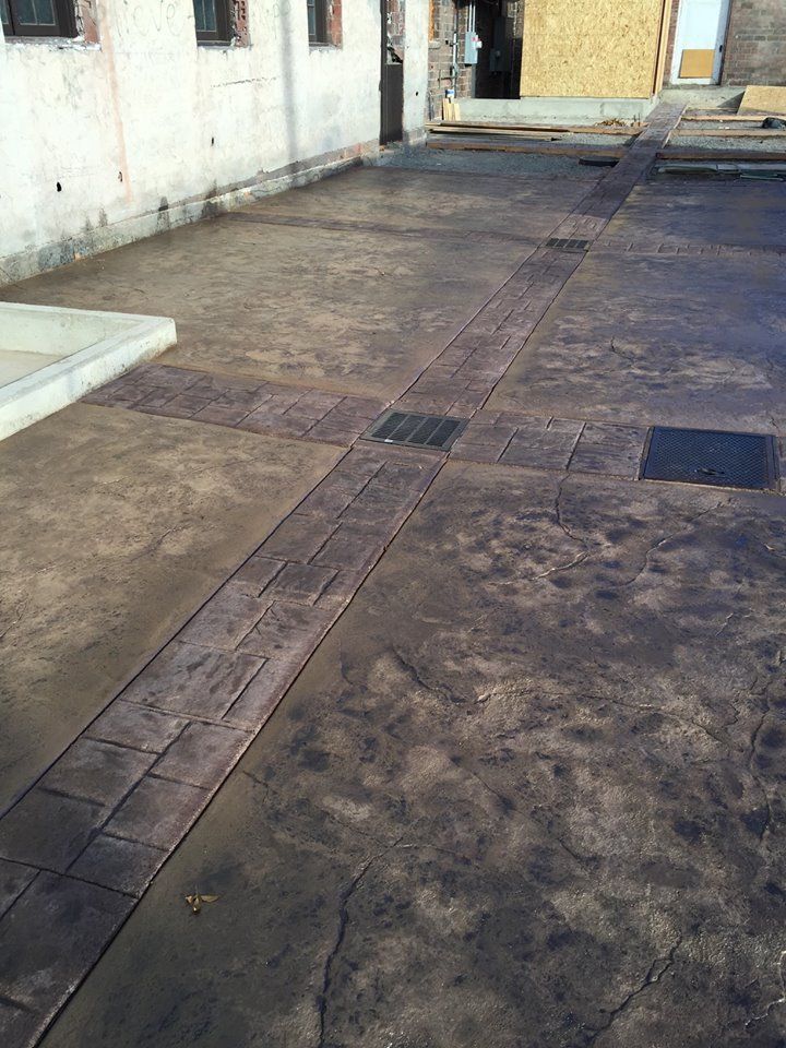 Outdoor paved walkway with dark concrete tiles and two square drain covers beside a white wall