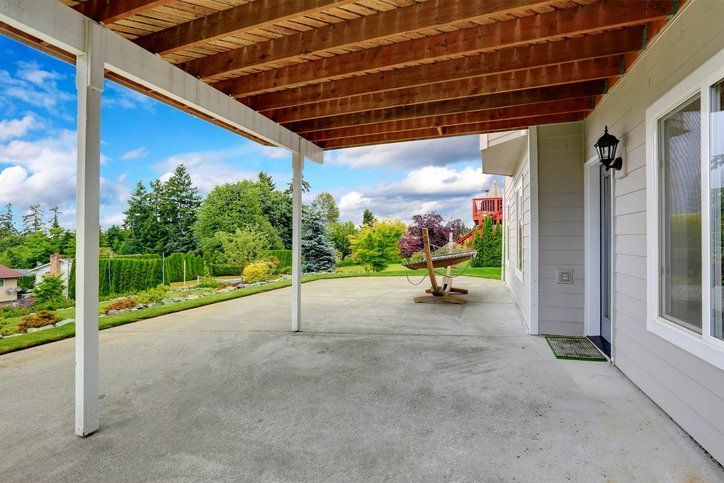 Covered concrete patio with exposed wood ceiling, white posts, and a view of a landscaped backyard.