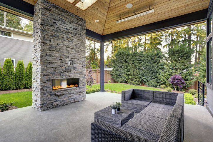 Covered outdoor patio with stone fireplace, gray wicker seating, and landscaped backyard