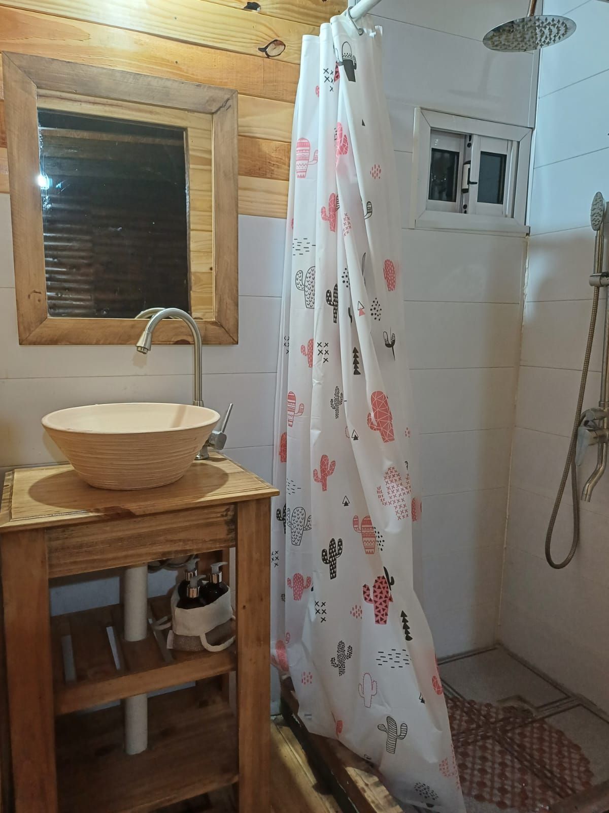 Baño con tocador de madera, lavabo tipo bol, ducha con cortina de cactus, ventana.
