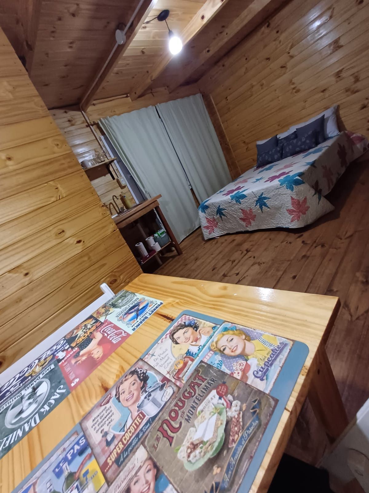 Dormitorio con paredes de madera, cama, mesa con anuncios antiguos, cortinas y mesita de noche.