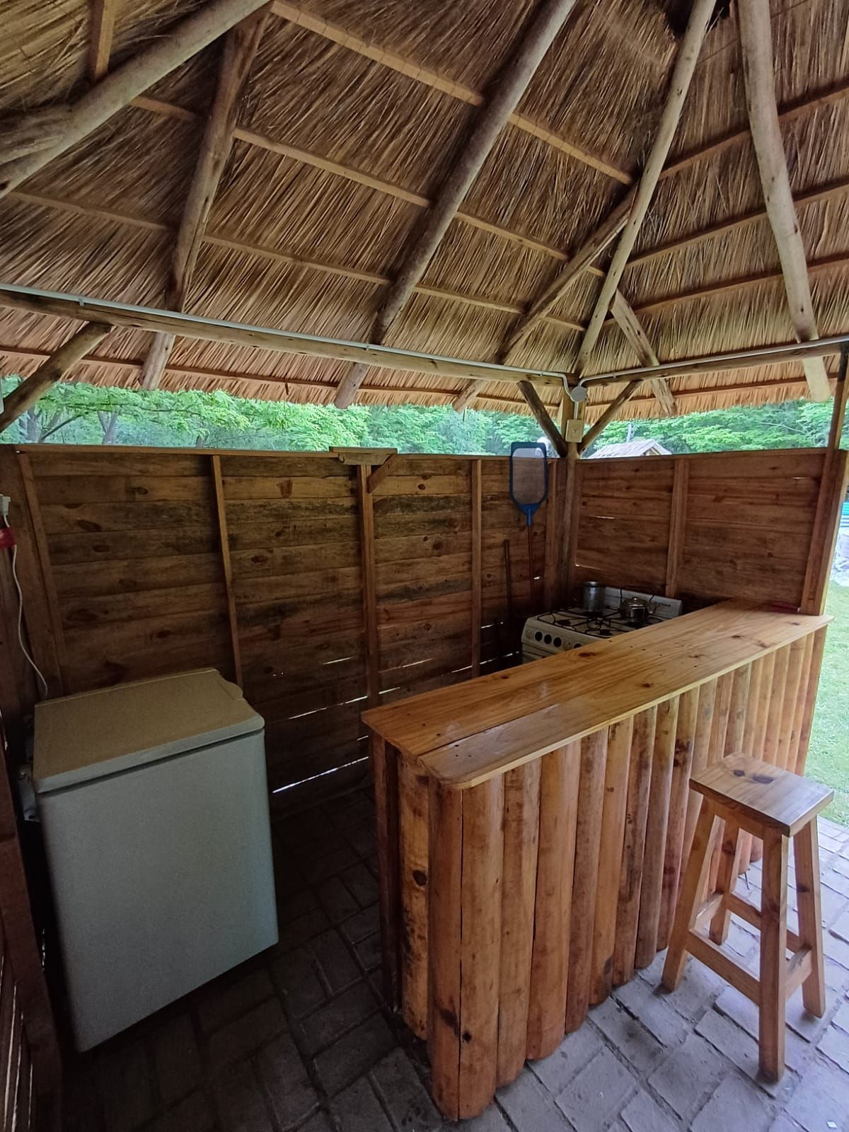 Bar exterior de madera bajo techo de paja, con nevera, taburete y pared trasera.