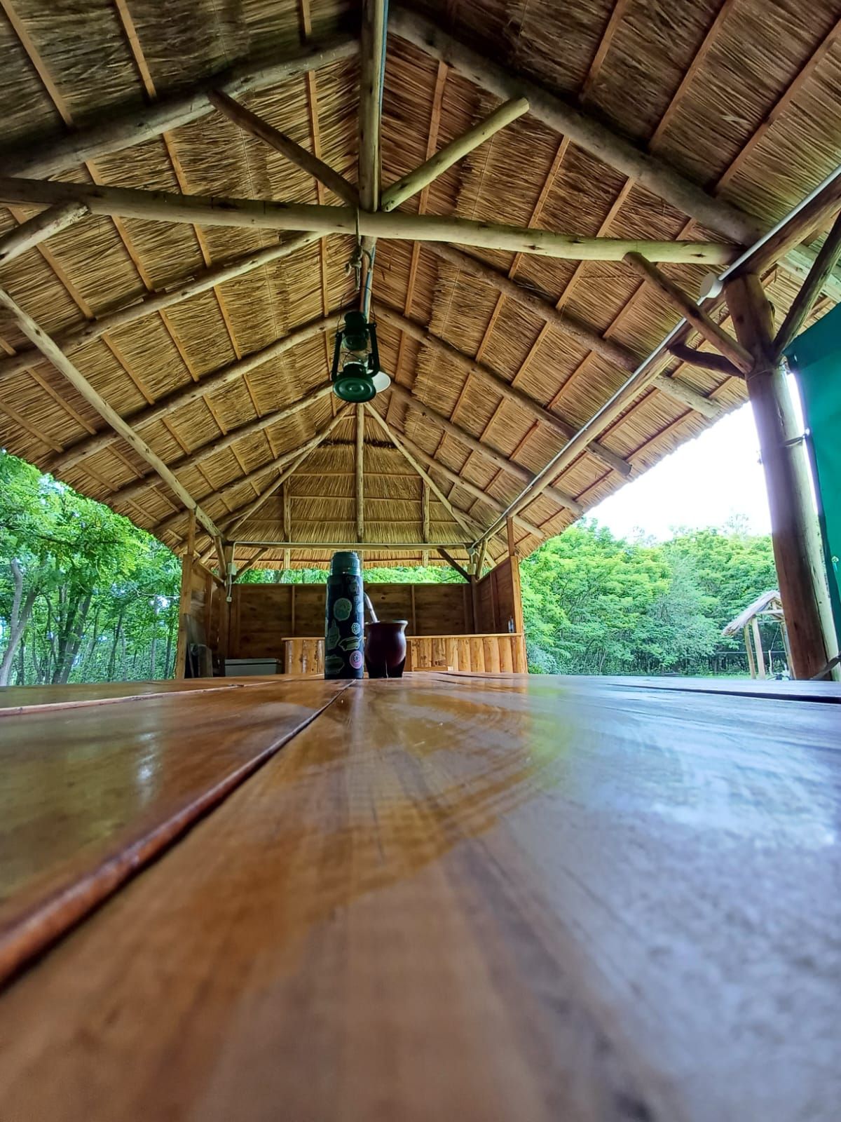 Glorieta de madera con techo de paja, con vistas a los árboles. En su interior cuelga una linterna y una mesa se extiende.