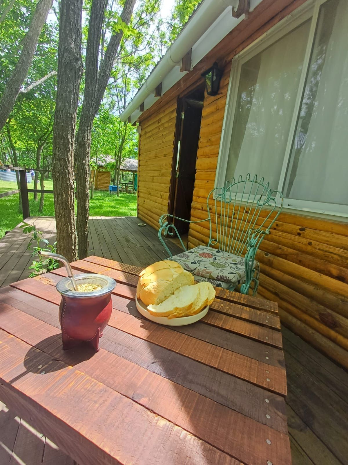 Exterior de una cabaña de madera con mesa puesta con pan y una bebida; silla verde; árboles.