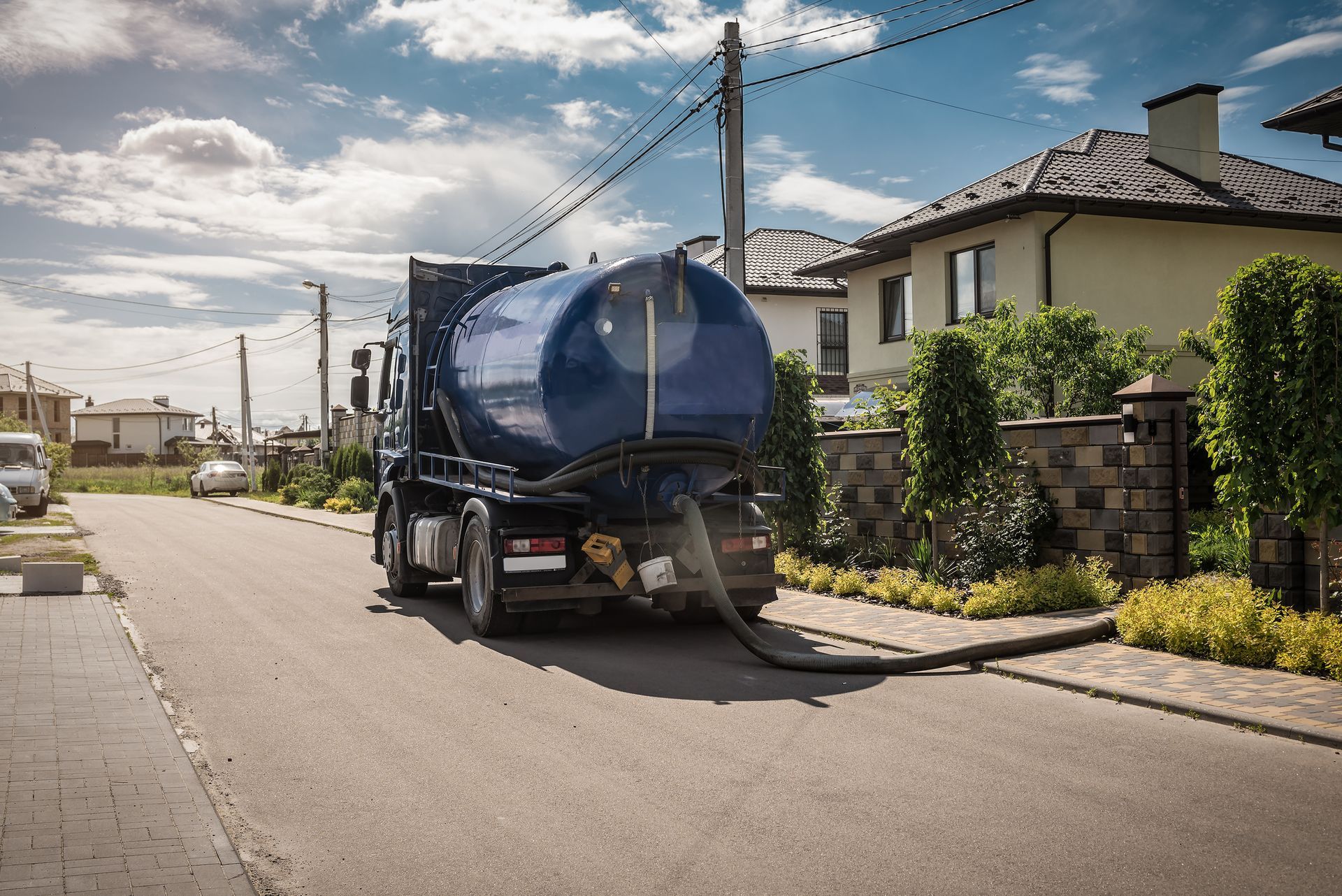 Septic tank truck parked on residential street, hose extended. Blue tank, sunny day.
