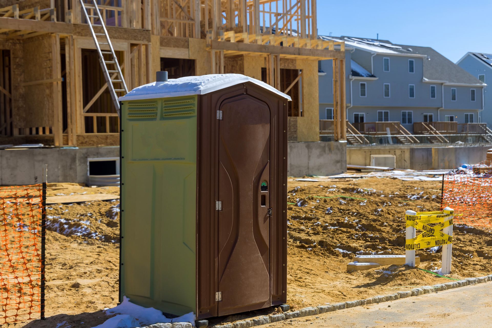 Brown and green portable toilet on a construction site with partially built houses in the background.
