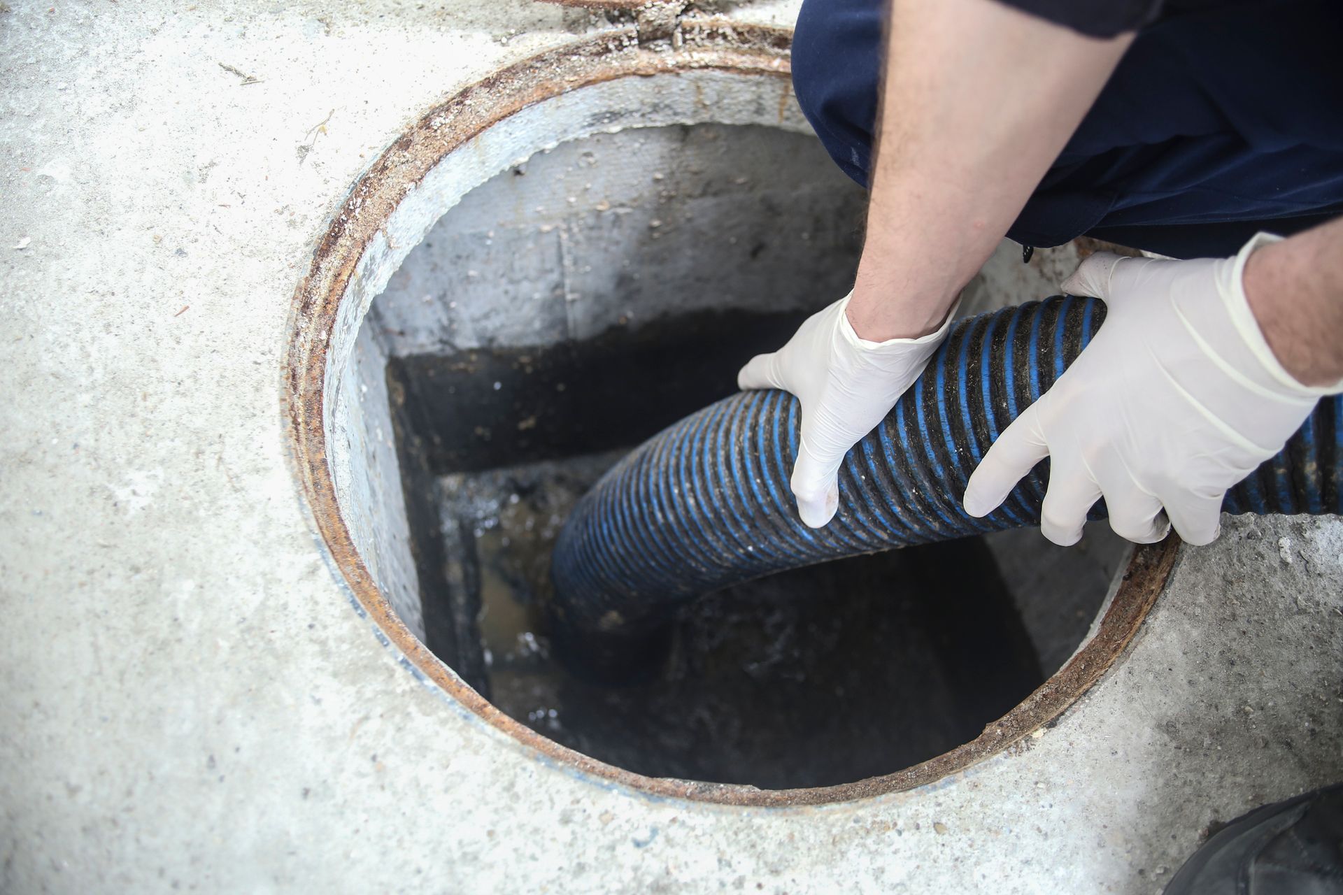 Person in gloves holding a hose into a dirty, open, concrete manhole.