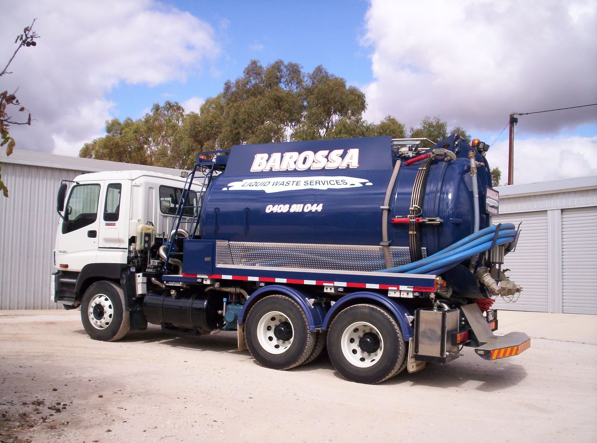 Blue and white Barossa septic tank truck parked on a gravel lot under a cloudy sky.