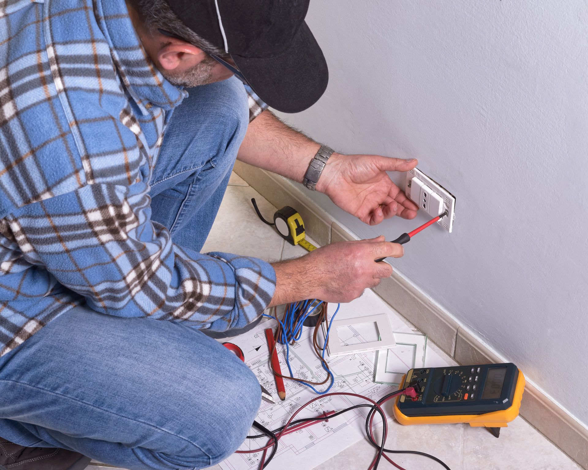 A man in a plaid shirt is kneeling on the floor working on an electrical outlet