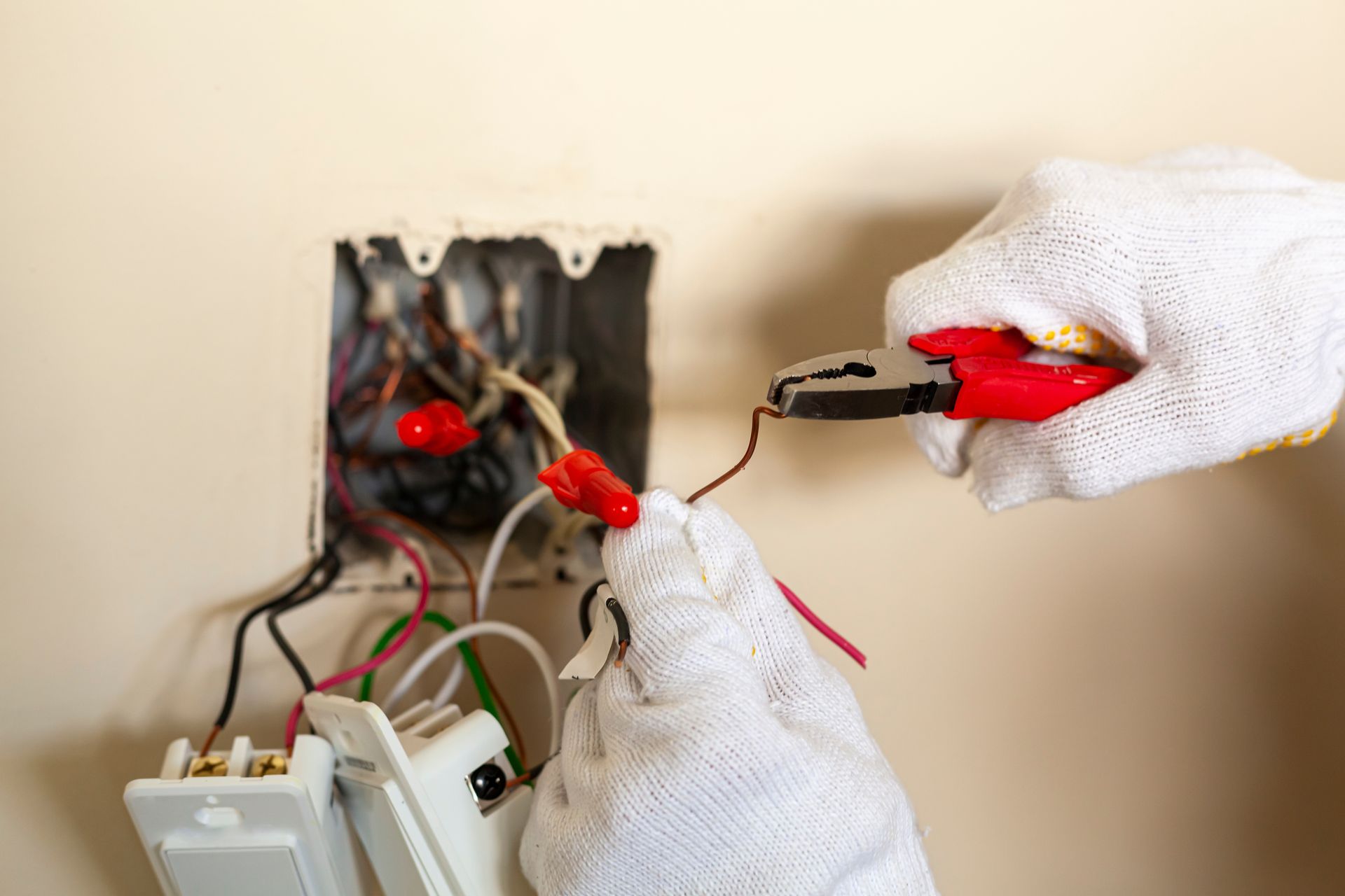 A person wearing gloves, working on electrical wiring in a wall outlet, using pliers.