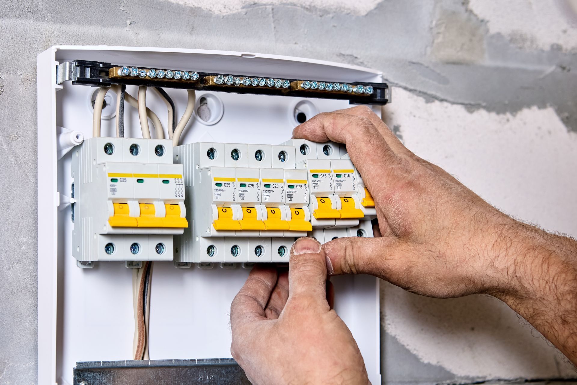 Hands installing circuit breakers in an electrical panel, with wires, on a concrete wall.