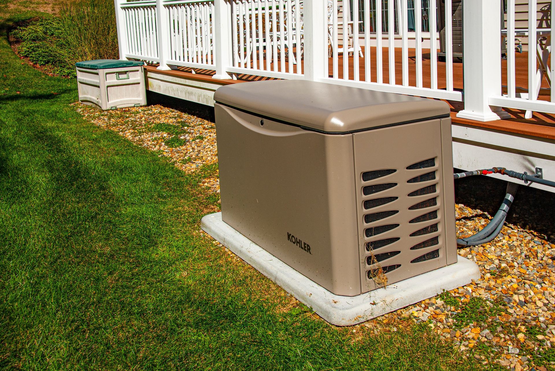 A tan Kohler backup generator sits on a concrete pad in a grassy yard next to a white wooden deck.