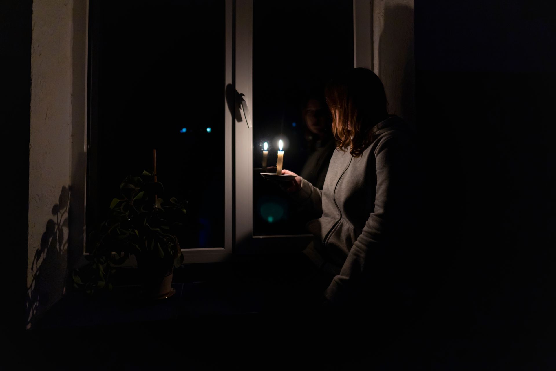Person holding a lit candle by a dark window, with another candle and a plant nearby.