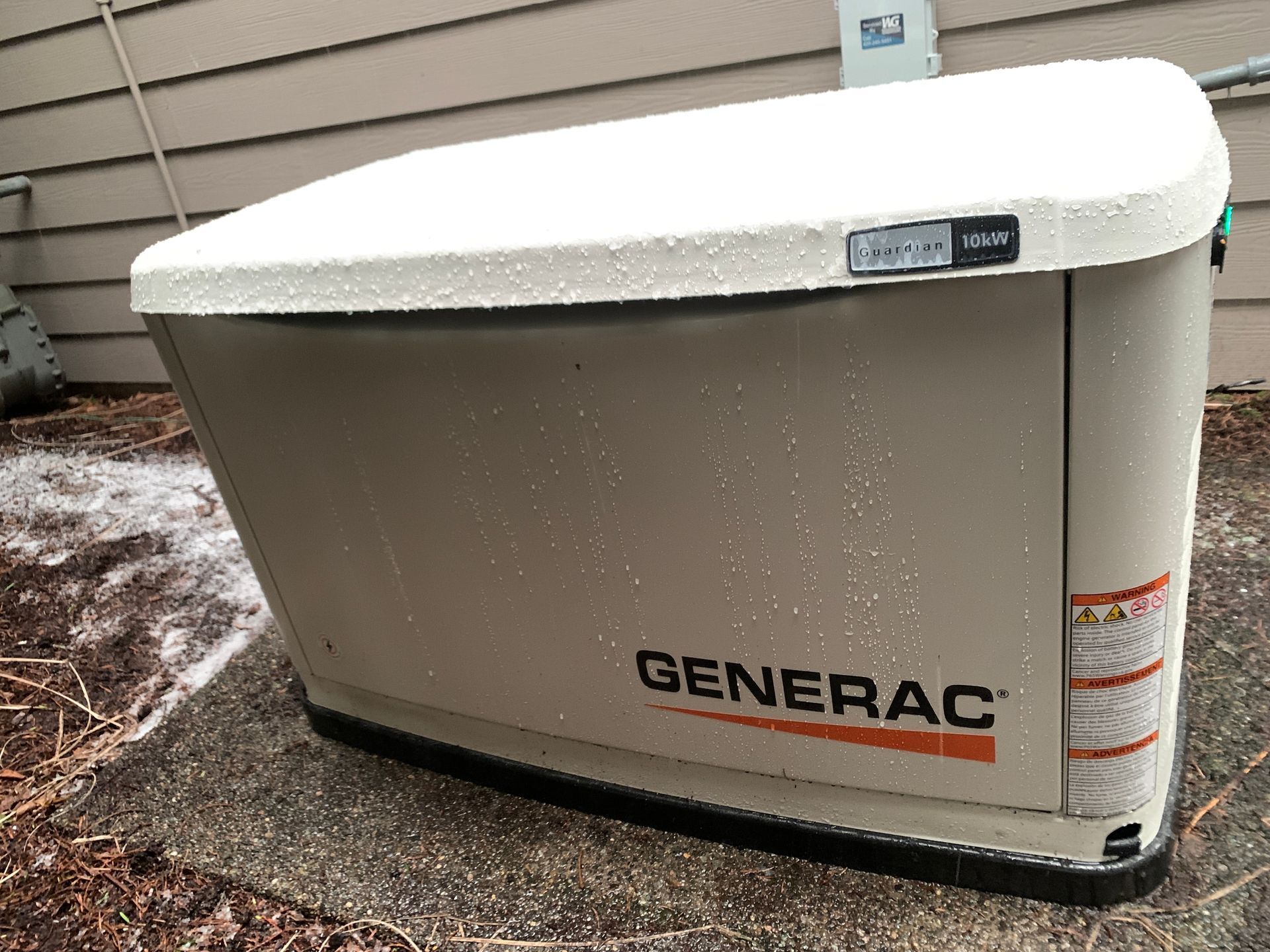 A tan Generac backup generator with a snowy lid sits on a concrete pad against the side of a house.