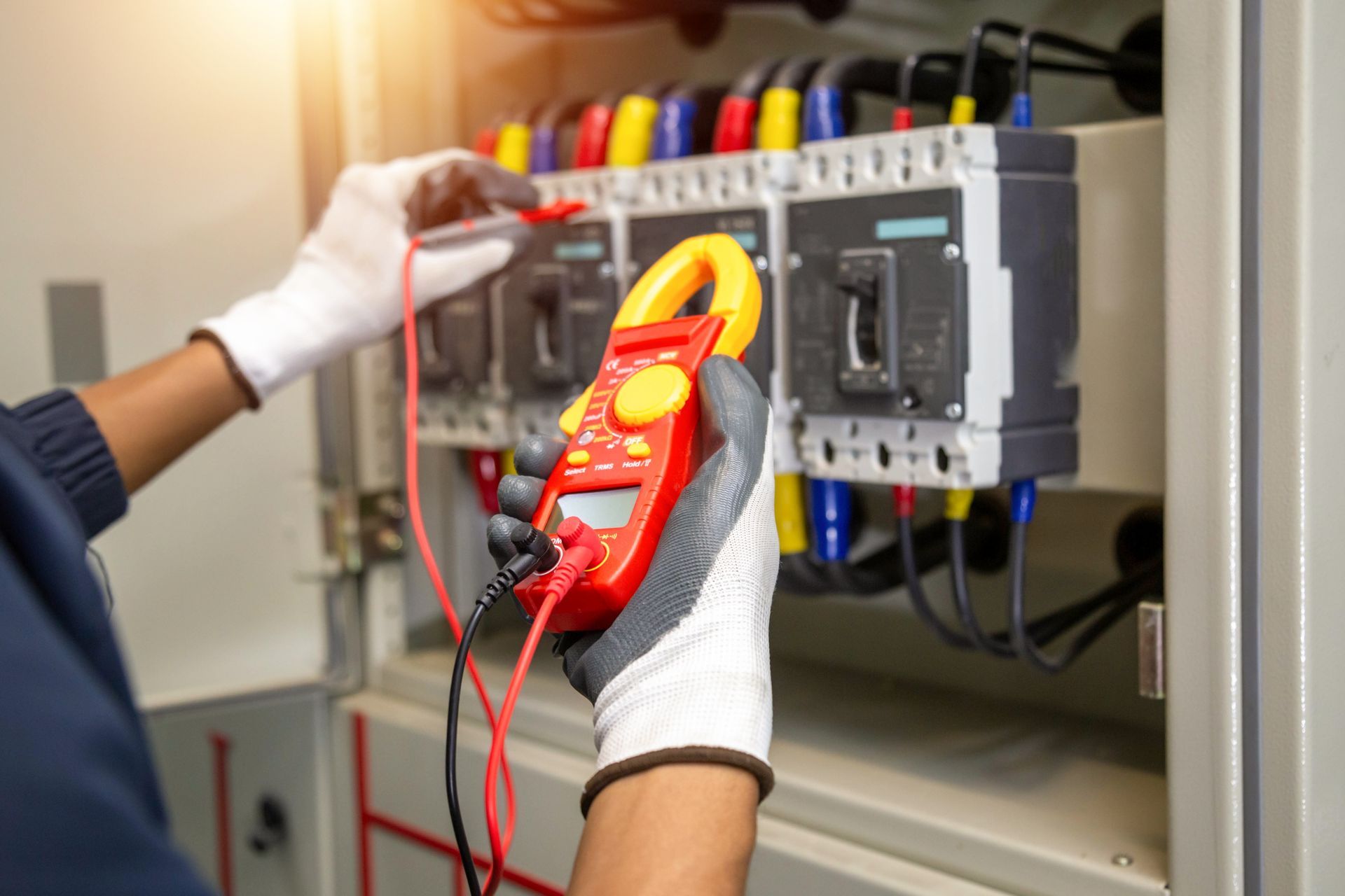 An electrician wearing gloves uses a red clamp meter to measure voltage at an electrical panel.