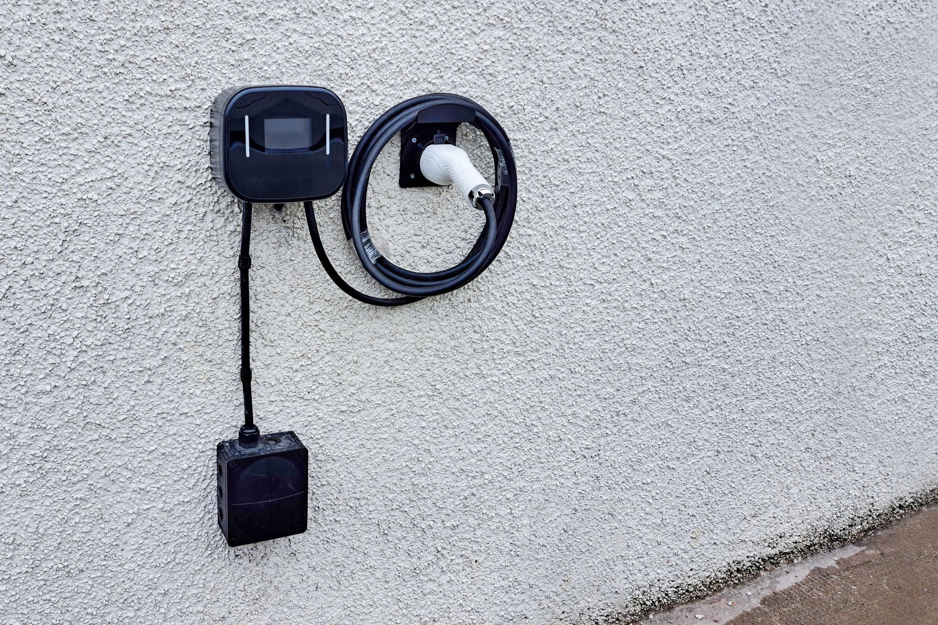 Black electric vehicle charger mounted on a textured, light gray wall, with a coiled cable and plug.