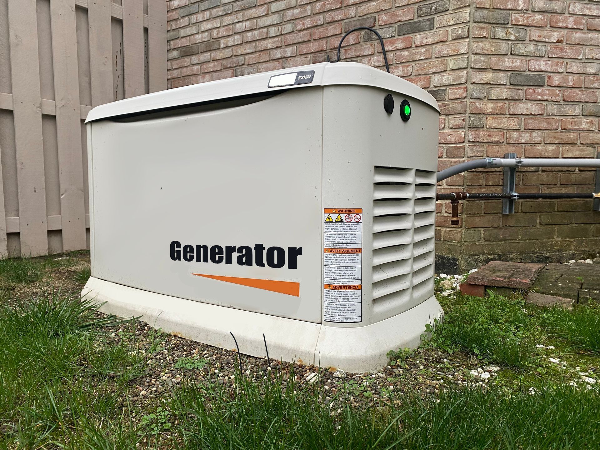White outdoor generator beside a brick building and grass, with ventilation vents and warning labels