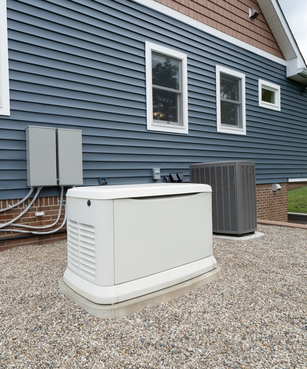A white home standby generator sits on a gravel pad outside a house with blue siding, next to an HVAC unit.