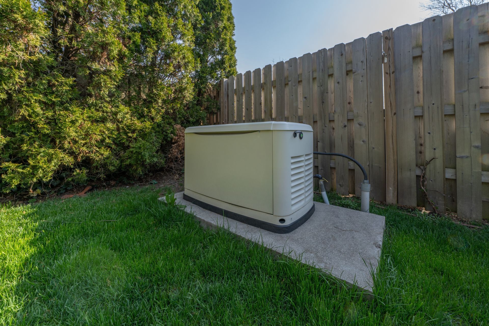 A beige backup power generator sits on a concrete pad in a grassy backyard, positioned next to a tall wooden fence.