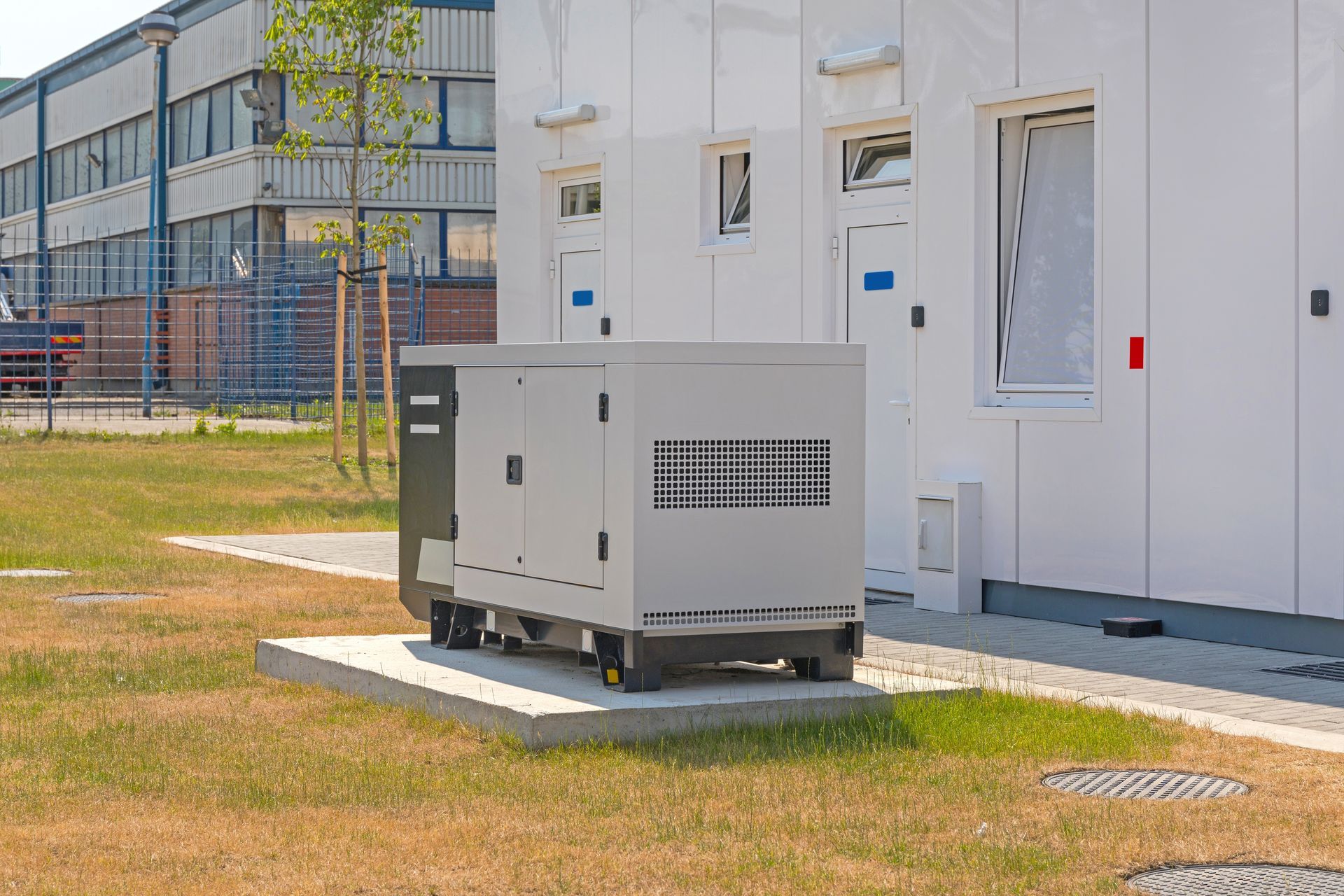 Gray industrial generator beside a white building on a grassy lot, with a modern building in the background