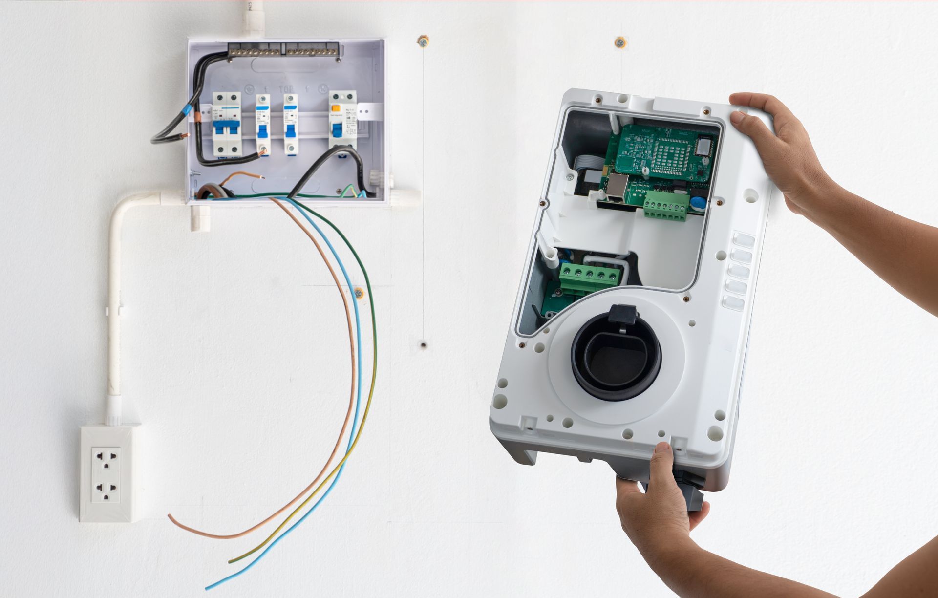 A person installs an electric vehicle wall charger next to an open electrical panel with exposed wiring on a white wall.