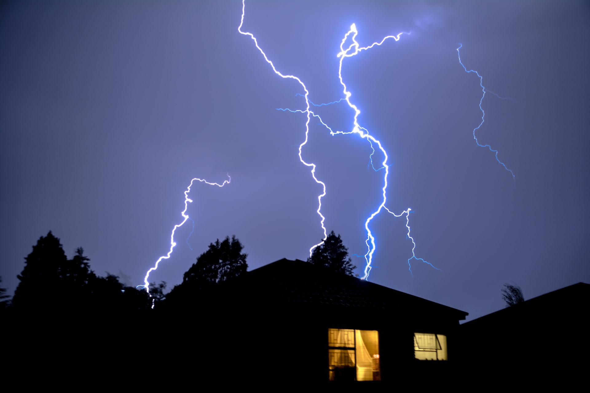 Lightning bolts over silhouetted rooftops at night, with a lit window in one house