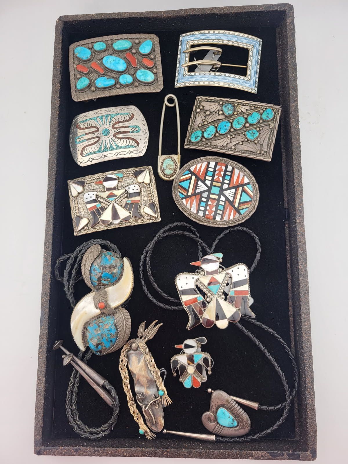 Jewelry display: silver and turquoise belt buckles, bolo ties, and a safety pin with turquoise and coral accents, arranged on black velvet.